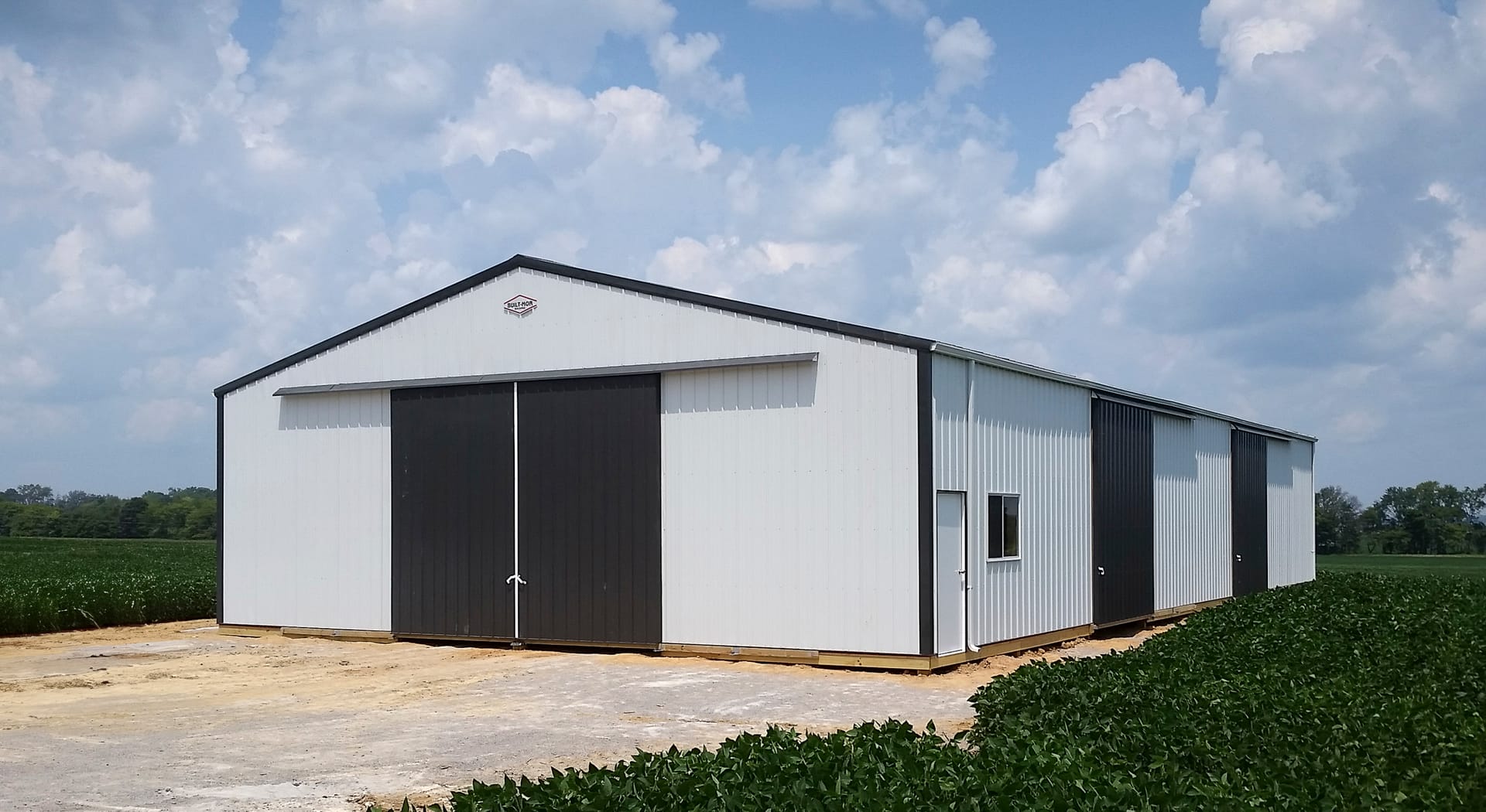 The image shows a large, rectangular post-frame building with white metal siding and a pitched roof, constructed by Built-Mor Buildings. It features contrasting dark sliding doors, suitable for vehicle or equipment entry. The building has at least three s