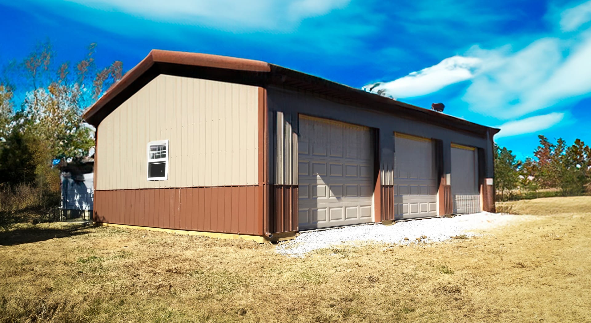 The image shows a rectangular post-frame building with a dual-tone exterior: light beige upper walls and dark brown lower siding and roof. It features three large garage doors, indicating its use as a workshop or storage facility. A small window is presen