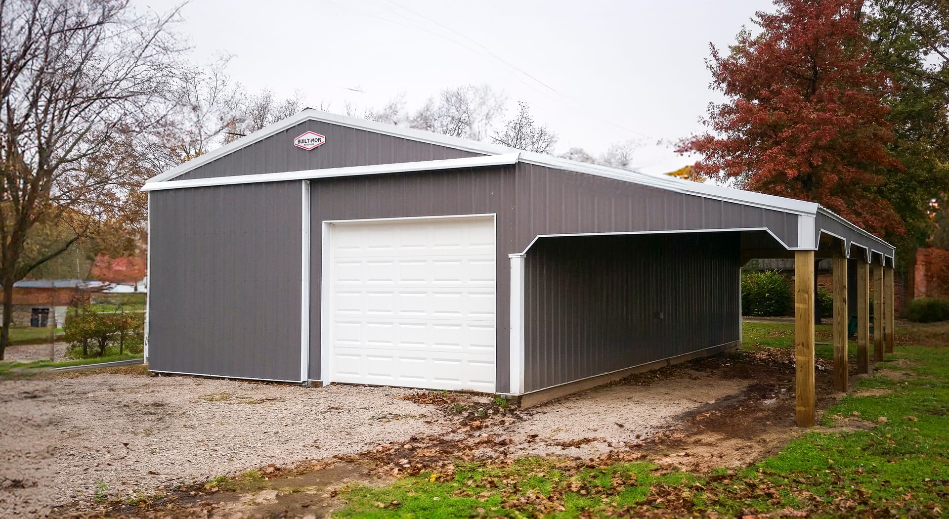 The image shows a post-frame building by Built-Mor Buildings, featuring a dark gray exterior with white trim. It includes a large white garage door, indicating its potential use as a garage or storage facility. The structure has a gable roof and an attach