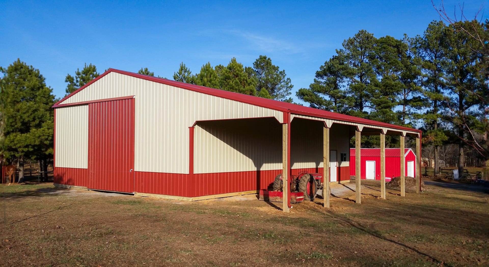 A post-frame building with red and beige vertical metal siding features prominently. It has a large sliding door on one end and an extended covered porch supported by wooden posts, indicative of agricultural or storage use. The roof is gently sloped and c
