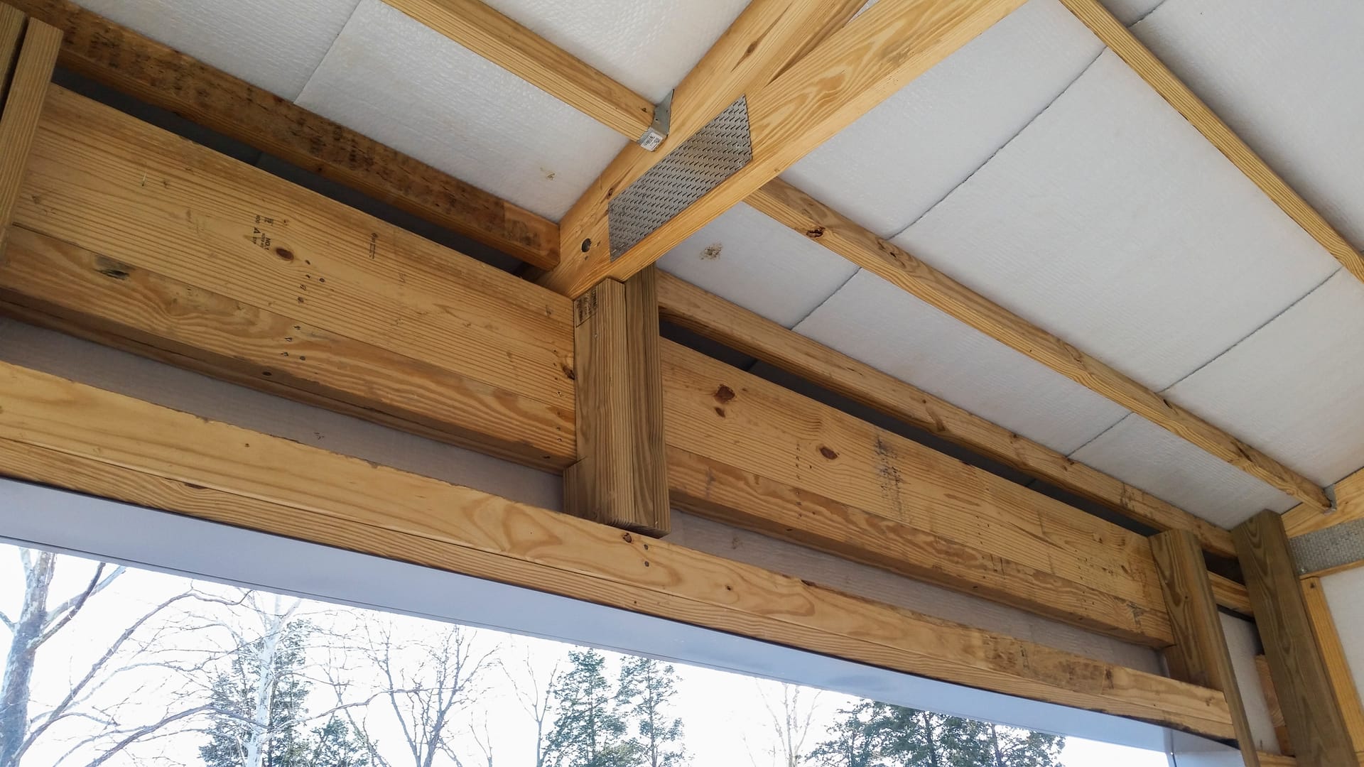 The image showcases a post-frame construction by Built-Mor Buildings, highlighting key structural elements such as wooden beams, posts, and trusses. The ceiling is insulated with a visible layer of white material, while metal fasteners secure the connecti