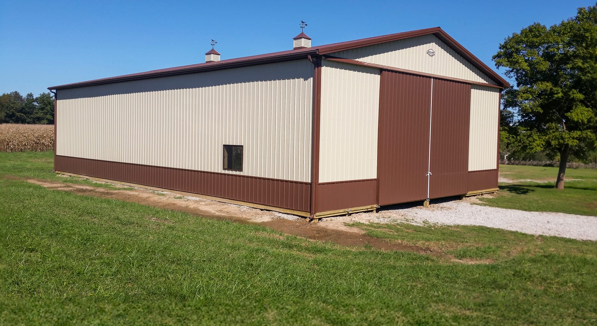 This image shows a post-frame building from Built-Mor Buildings, featuring a rectangular design with beige and brown vertical siding. The structure has a gabled roof with two cupolas. A large sliding door is located at the front, and a small window is pre