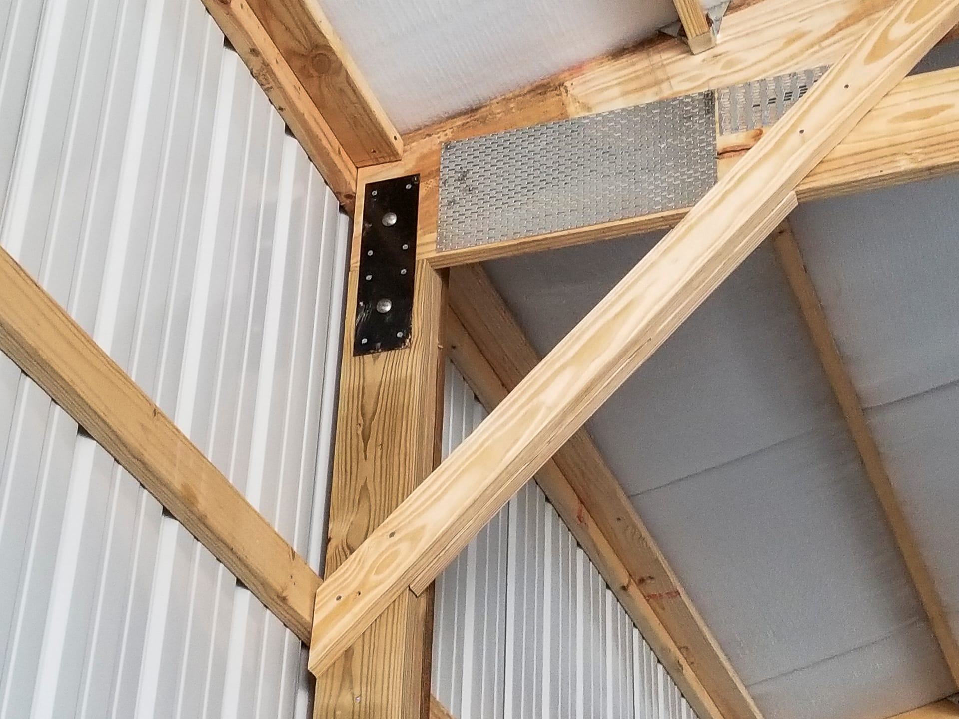 The image shows a detailed view of a post-frame construction corner from Built-Mor Buildings. It features wooden beams with metal connectors and braces for structural support. The interior wall is lined with vertical white metal panels, and insulation is