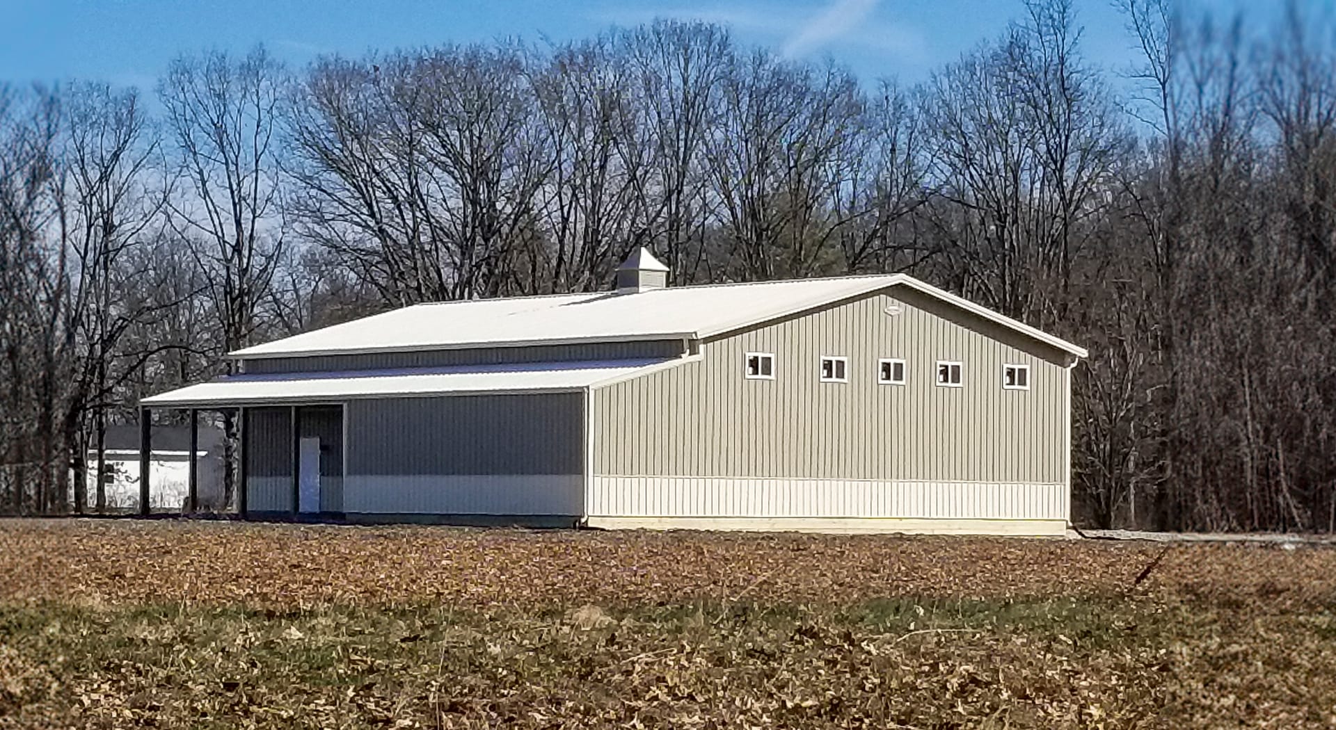 The image features a post-frame construction building from Built-Mor Buildings, designed for agricultural purposes. It has a light gray exterior with white trim and a gabled roof. There are small square windows along the side, and a large sliding door at