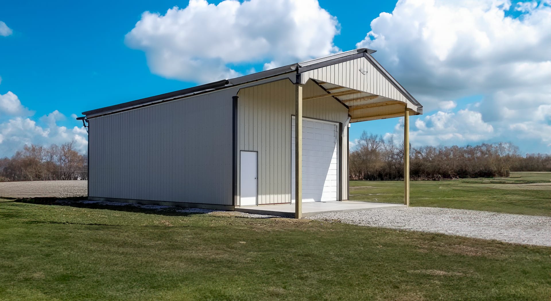 This image shows a post-frame building with beige metal siding and a gabled roof, featuring a large roll-up garage door at the front. The structure includes a small adjacent side door and an extended covered porch supported by two vertical posts. The buil