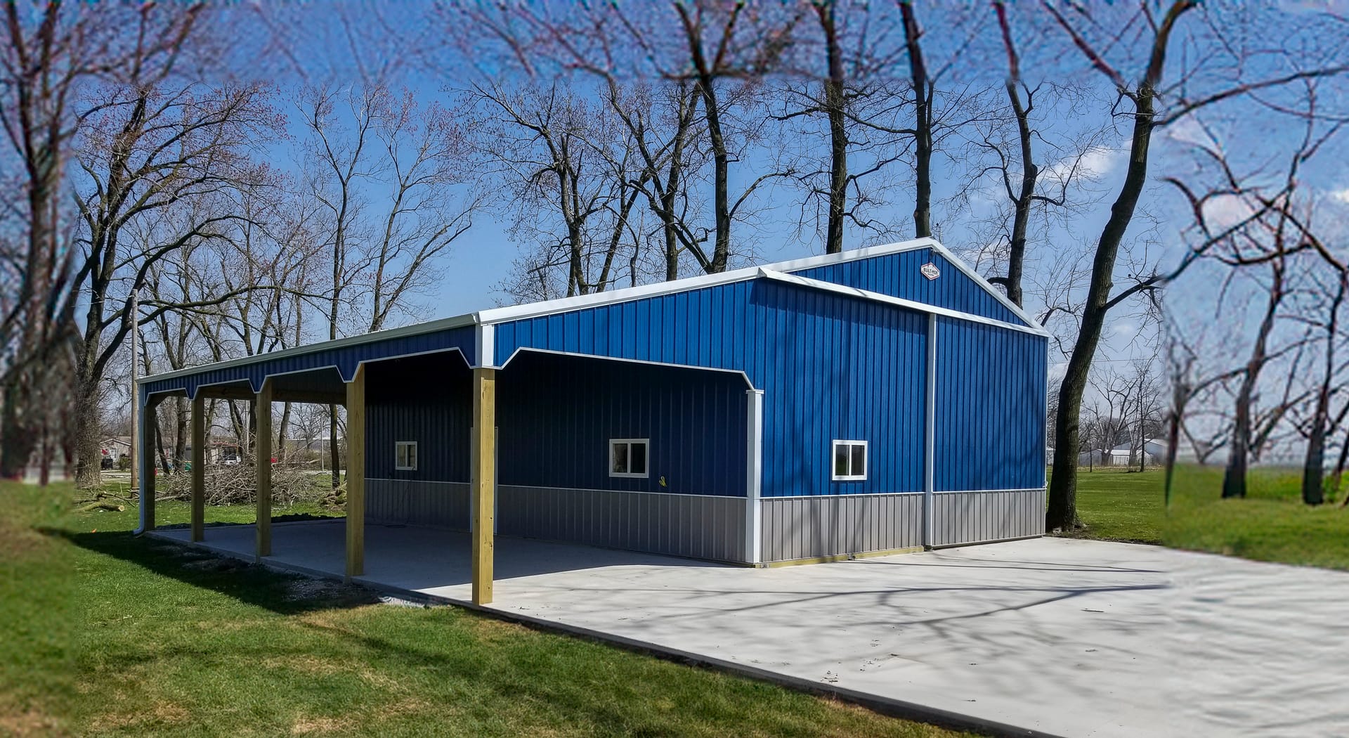 A blue post-frame building by Built-Mor Buildings features a gabled roof and a combination of metal siding in blue and light gray. The structure includes a covered open area supported by wooden posts, likely used as a carport or patio. The building is sit