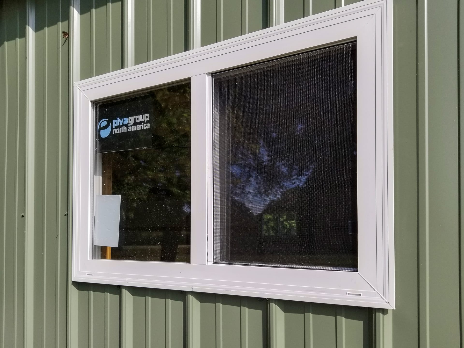 Close-up image of a post-frame building featuring a green metal siding with vertical ridges. A white-framed rectangular window is centrally positioned, with one section containing a mesh screen. Inside the window, signage for “pivagroup north america” is