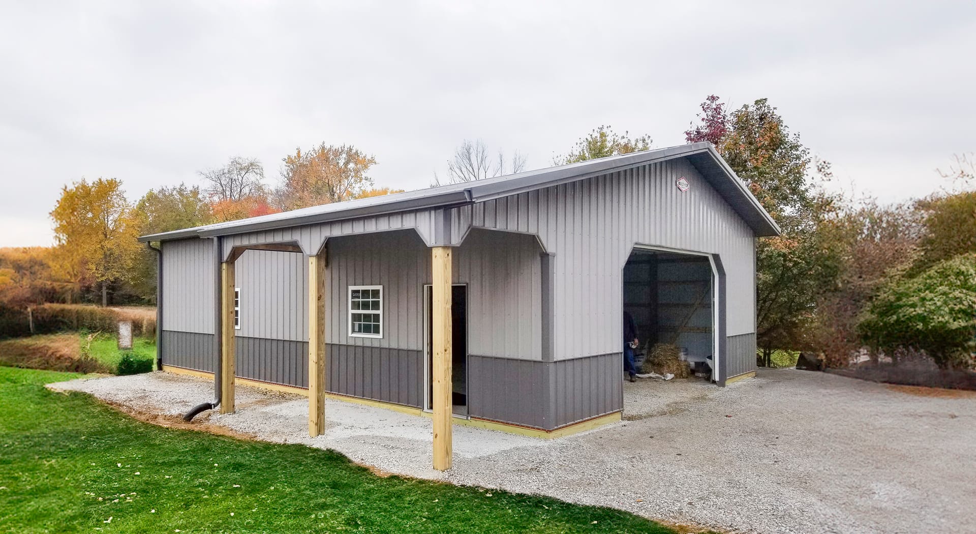 The image depicts a post-frame construction building by Built-Mor Buildings, primarily made of steel with a gabled roof. The structure features a large open bay on one side, indicating its use as a storage or workspace, likely a garage or workshop. It has