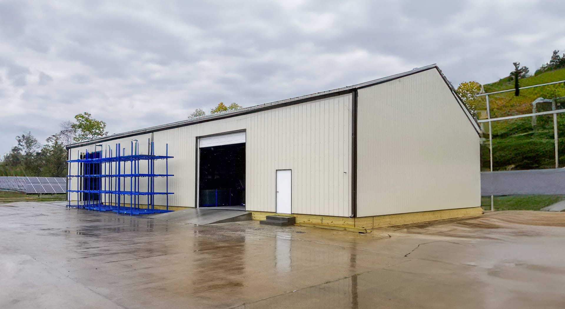 The image depicts a post-frame construction building designed for industrial or storage purposes. The structure features a peaked roof and light-colored metal siding. It includes a large, open sliding door for vehicle or equipment access and a smaller per