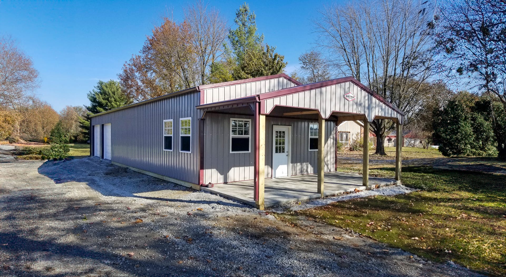 The image shows a post-frame building from Built-Mor Buildings, featuring metal siding and roofing with light gray and maroon trim. It has a gable-style roof and an extended entrance porch supported by wooden posts. The structure includes multiple windows