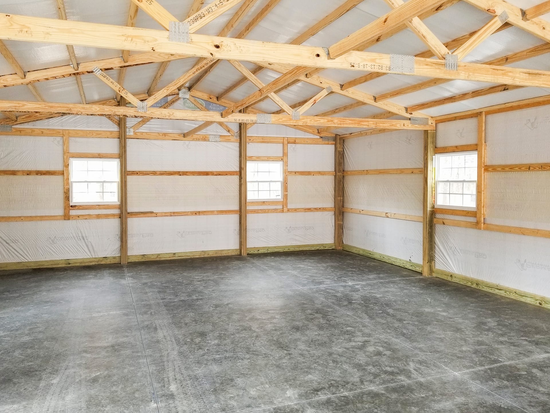 Interior of a post-frame building by Built-Mor Buildings, featuring exposed wooden trusses and beams supporting a pitched roof. The walls are lined with insulation panels and framed with horizontal wood members. The floor is concrete, suggesting use for s