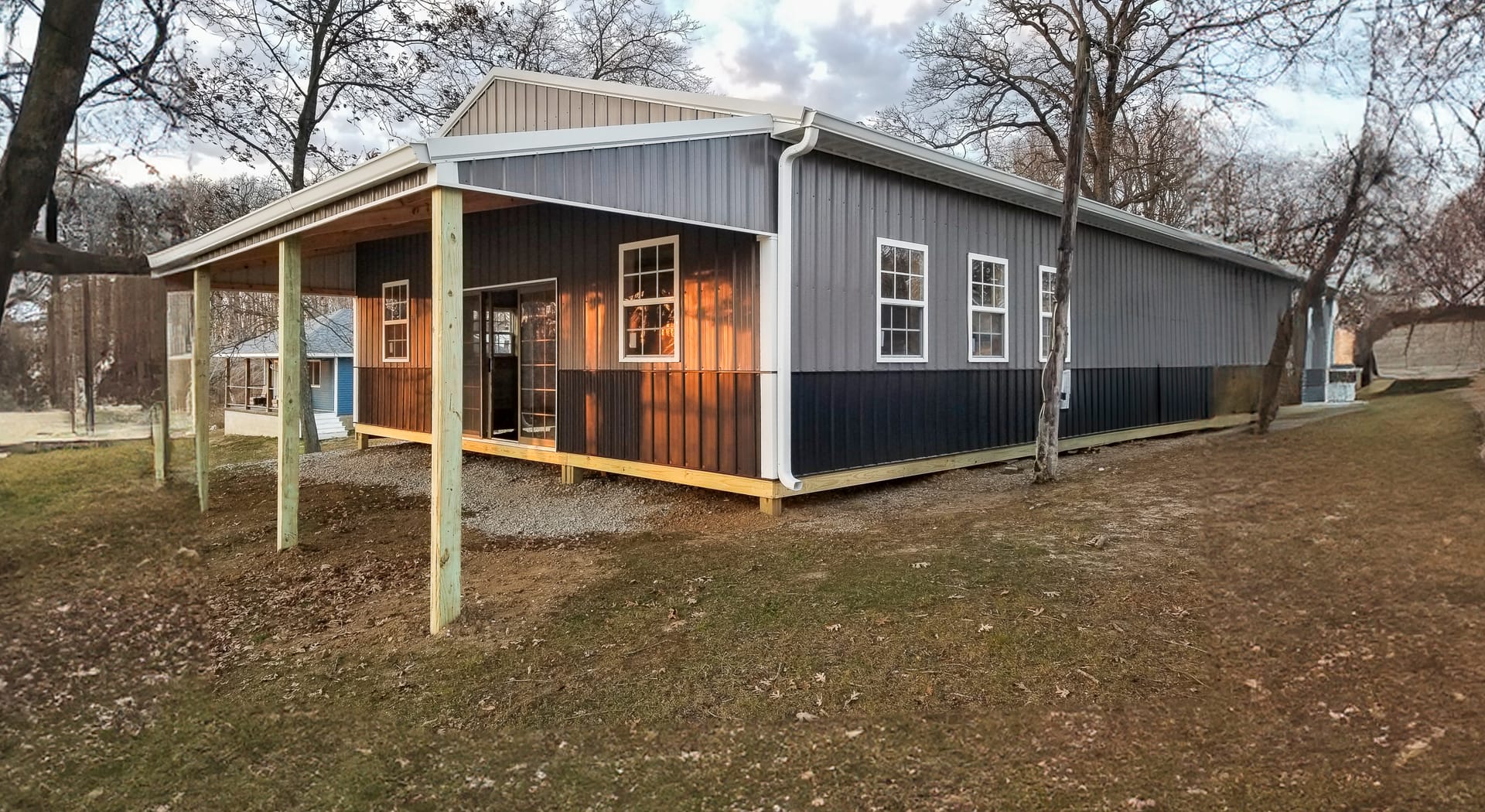 The image shows a post-frame construction building from Built-Mor Buildings. It features a gable roof and an extended covered porch area supported by wooden posts. The exterior walls have a two-tone design with vertical siding, light gray on the upper hal