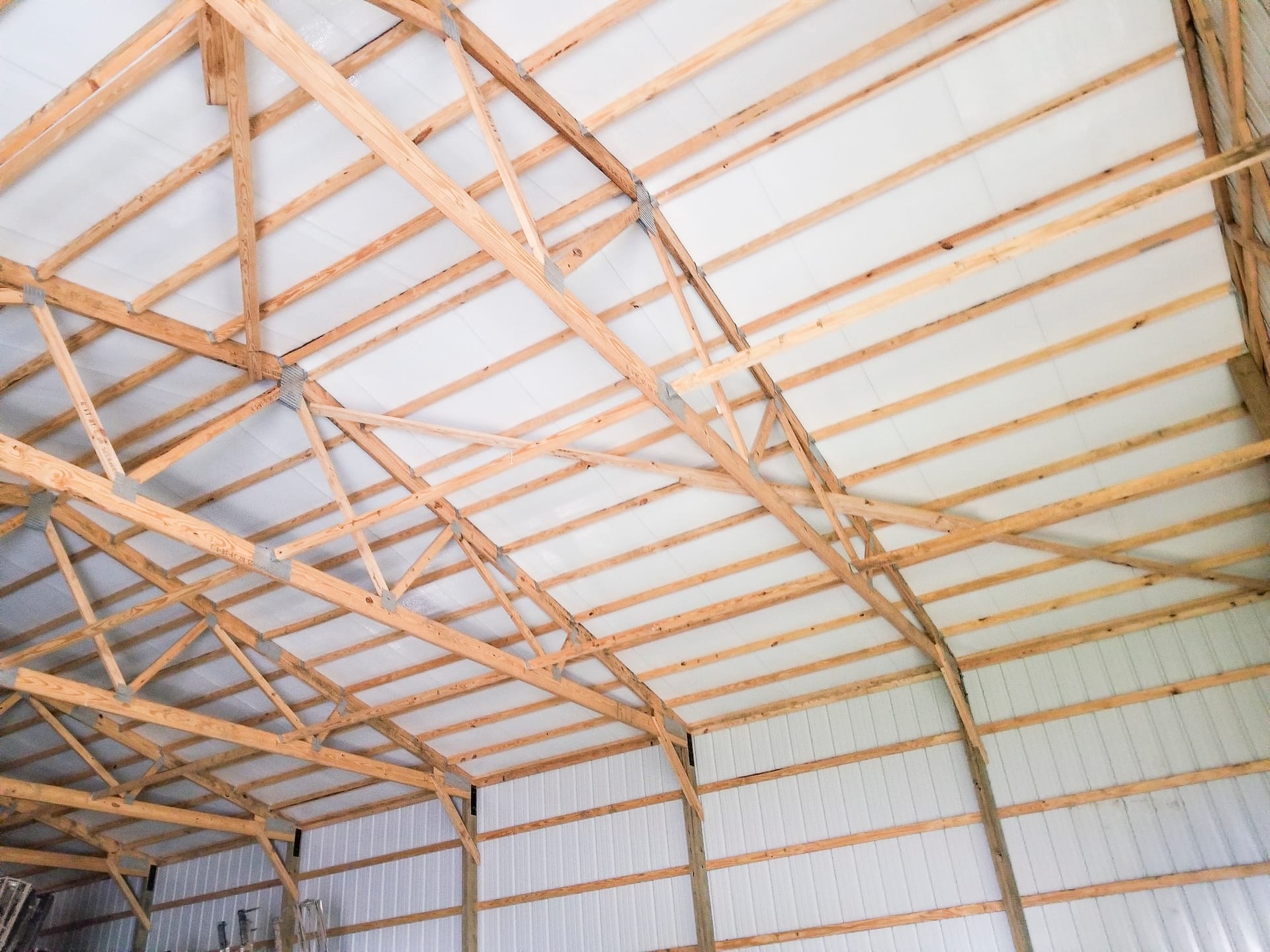 This image shows the interior of a post-frame construction building by Built-Mor Buildings. The structure features wooden trusses with metal connectors supporting a white ceiling. The walls are lined with vertical metal panels. The trusses are arranged in