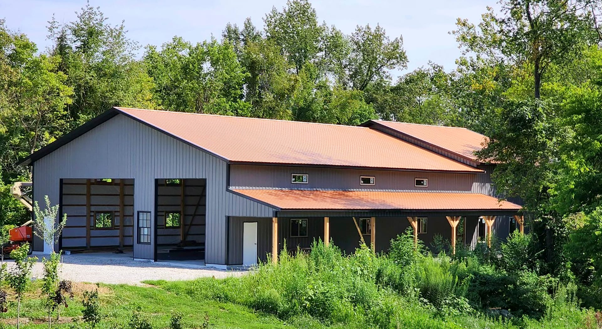 The image shows a post-frame building by Built-Mor Buildings, featuring a large, rectangular structure with a pitched metal roof. The building has two distinct sections: a taller main area with large open bays, likely for storage or workspace, and a lower