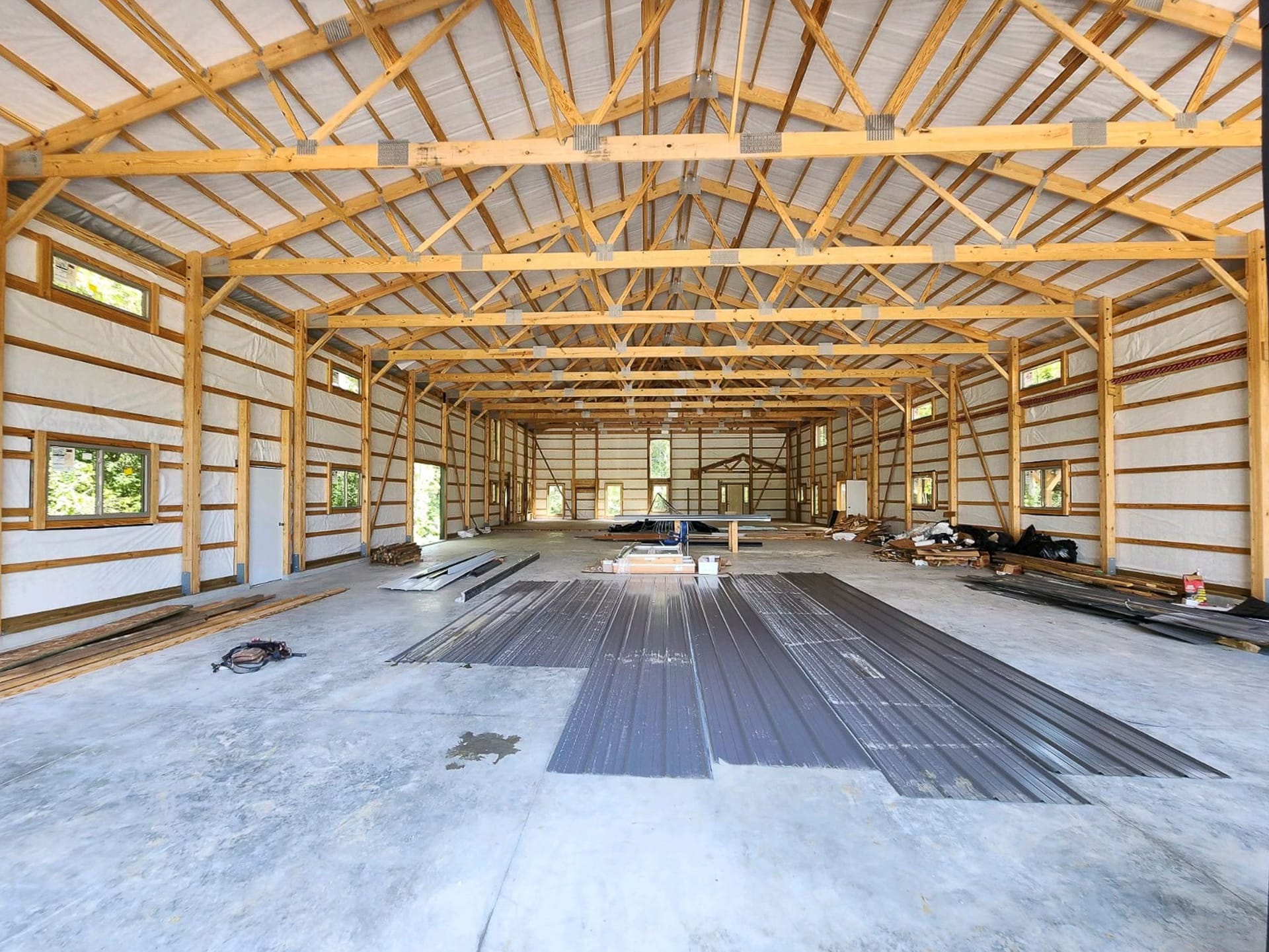 Interior view of a post-frame construction building by Built-Mor Buildings, featuring a spacious, open layout with exposed wooden trusses supporting the roof. The structure has insulated walls and large, evenly spaced windows allowing natural light. Build