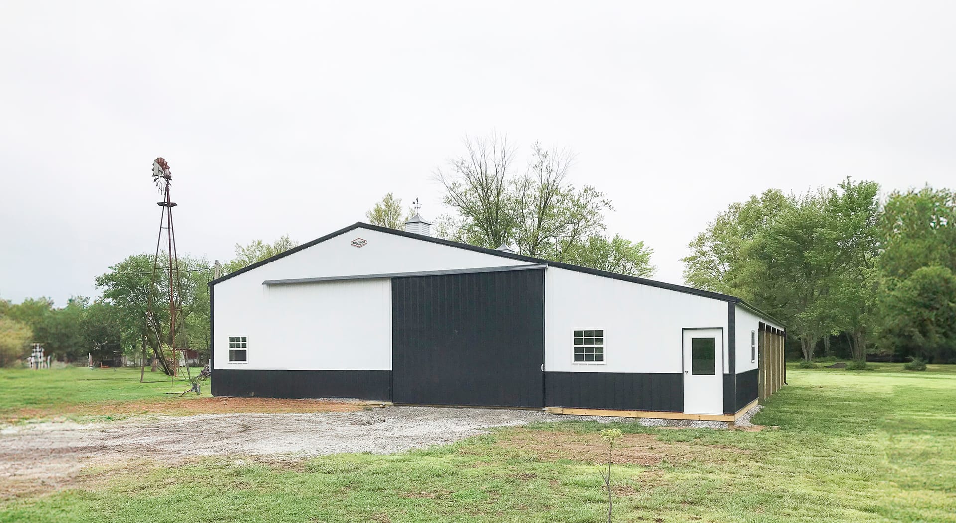 A post-frame construction building by Built-Mor Buildings is shown in a rural setting. The structure has a classic barn-like design with a gable roof and large sliding doors on the front facade. It features a two-tone color scheme with white upper walls a