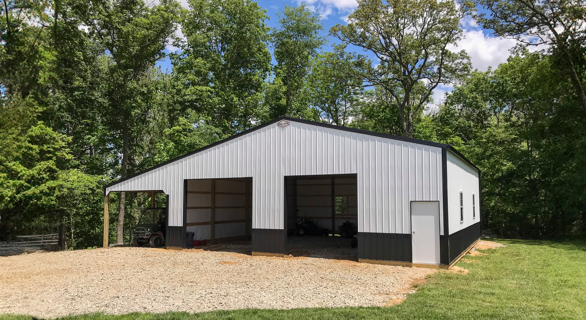 This image shows a post-frame building from Built-Mor Buildings, set against a wooded background. The structure features a white exterior with dark lower trim and includes three large open bays and a single-entry door, suggesting use as a garage or storag