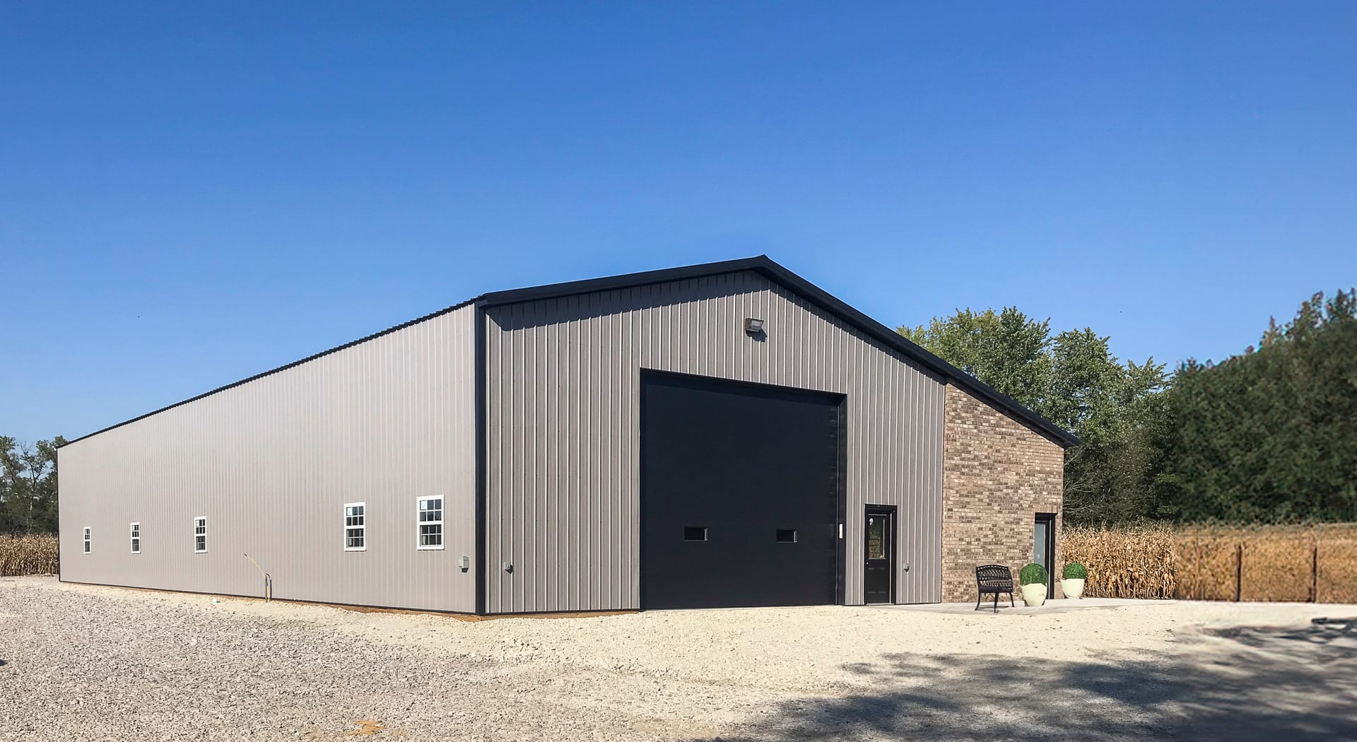 The image depicts a large post-frame building from Built-Mor Buildings, featuring a steel exterior and a gable roof. The front facade has a large black roll-up door and several white-framed windows. An entry door is adjacent to the roll-up door. The struc