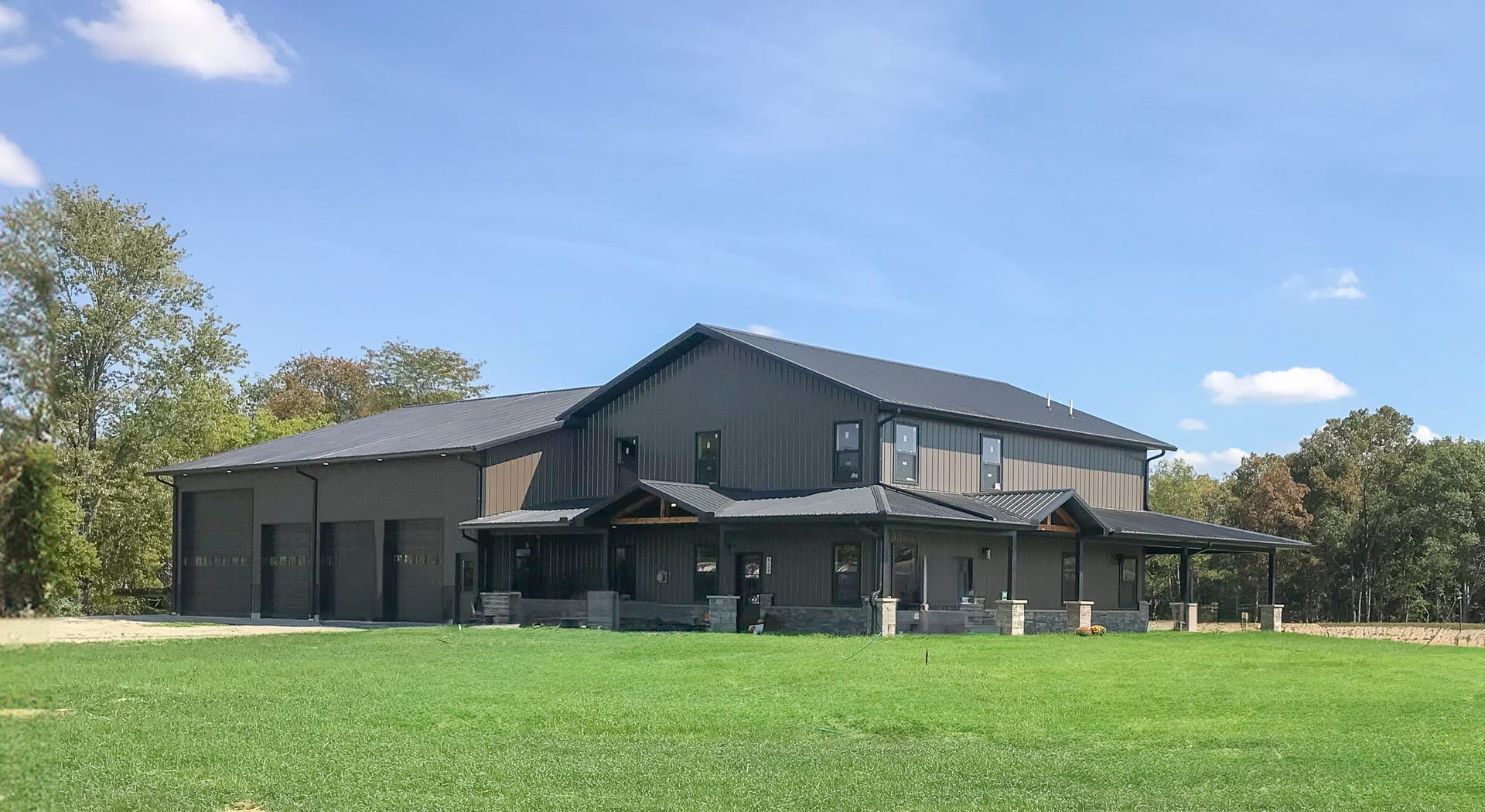 A large post-frame construction building with a dual purpose: part residential and part garage. The structure features a dark metal exterior with multiple sloped roofs. The residential section has several windows and an extended porch with stone pillars.