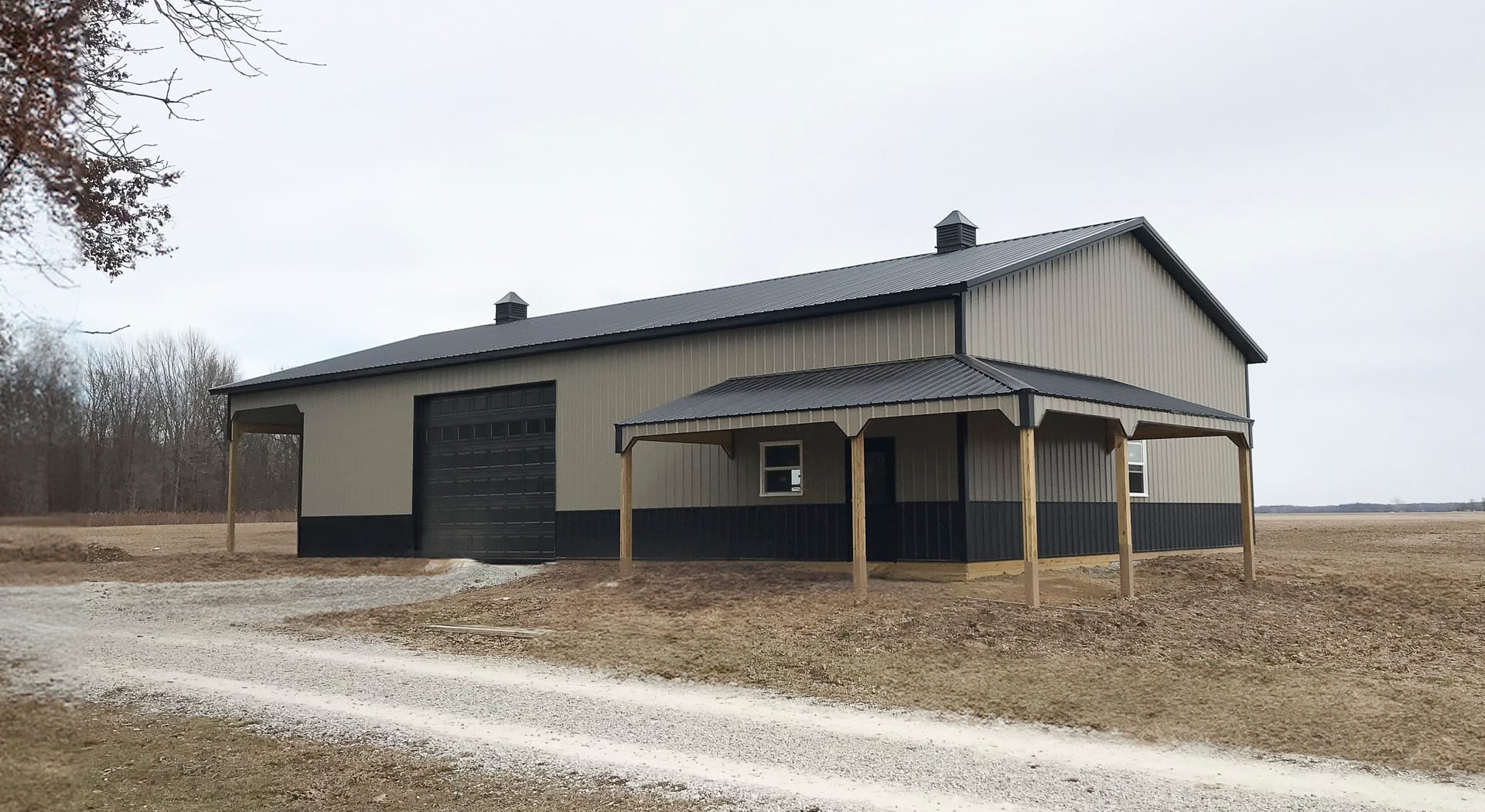 A post-frame construction building by Built-Mor Buildings is shown in a rural setting. The structure features a gable roof with two cupolas for ventilation and a combination of light and dark siding for contrast. A large overhead garage door suggests use